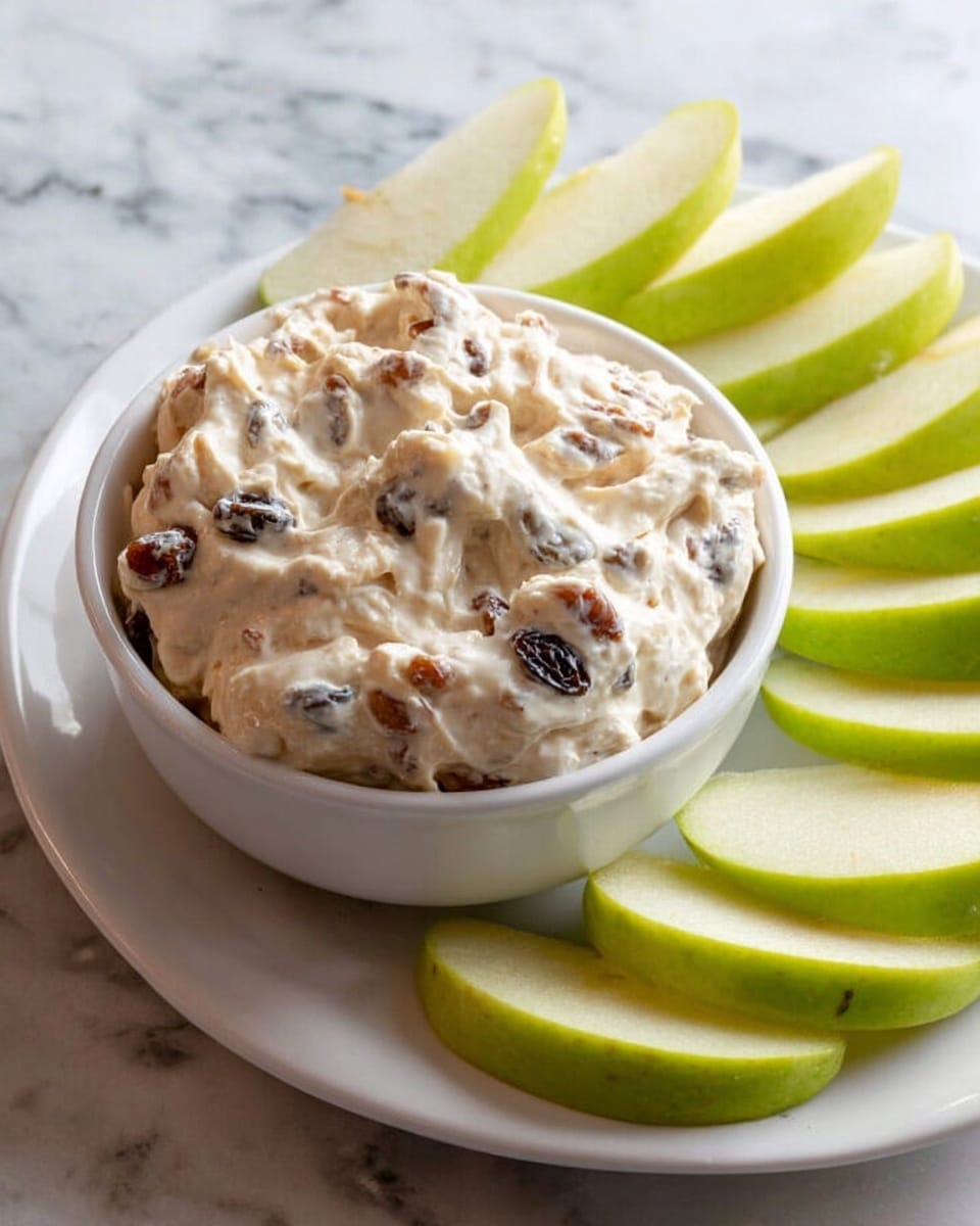 A small white bowl filled with a thick, creamy dip that is light beige with visible dark raisins and small brown nut pieces mixed throughout. The bowl sits on a white plate surrounded by bright green apple slices with their skin on, arranged close together but not overlapping. The background features a white marbled texture. photo taken with an iphone --ar 4:5 --v 7