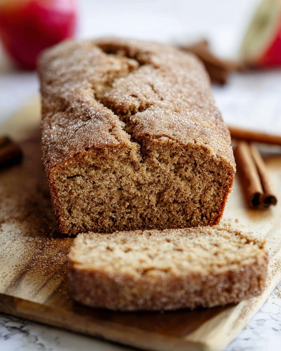 The image shows a loaf of brown bread with a crumbly texture, sliced to reveal a soft, dense inside. The crust is sprinkled with a layer of sugar, giving it a slightly rough and sparkling look. The loaf rests on a wooden board, and the visible slice lies flat in front of the loaf. In the background, a red apple and cinnamon sticks can be seen blurred out. The whole scene is set against a white marbled surface. photo taken with an iphone --ar 4:5 --v 7