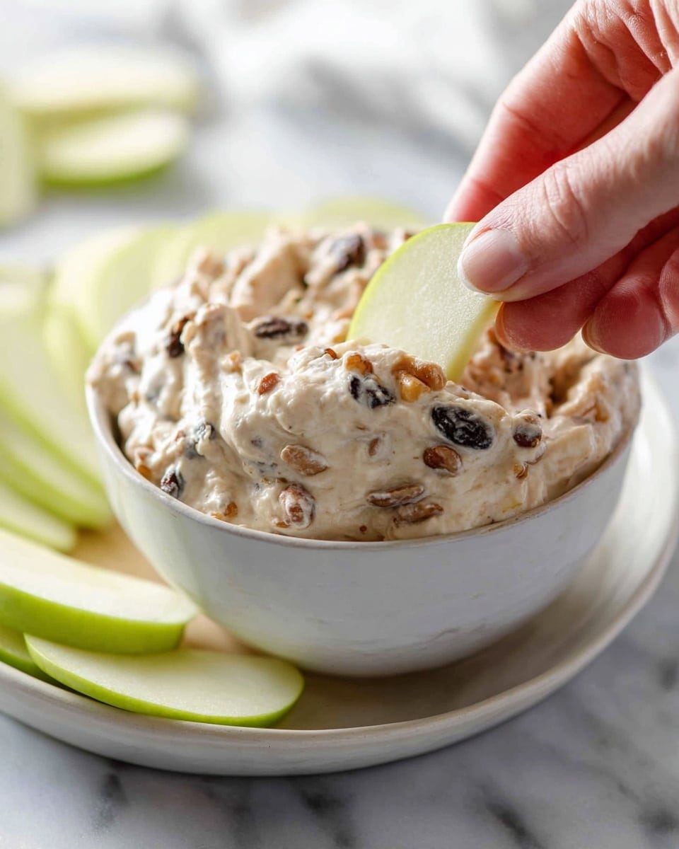 A close-up view shows a small white bowl filled with a creamy mixture that has visible nuts and dark raisins mixed throughout. A woman's hand is dipping a thin, light green apple slice into the thick, textured dip. The creamy dip is light beige in color with a rough texture from the nuts and raisins, and the apple slice is smooth with a slight sheen. The bowl sits on a matching white plate on a white marbled surface, with a few more apple slices scattered nearby in soft focus. photo taken with an iphone --ar 4:5 --v 7