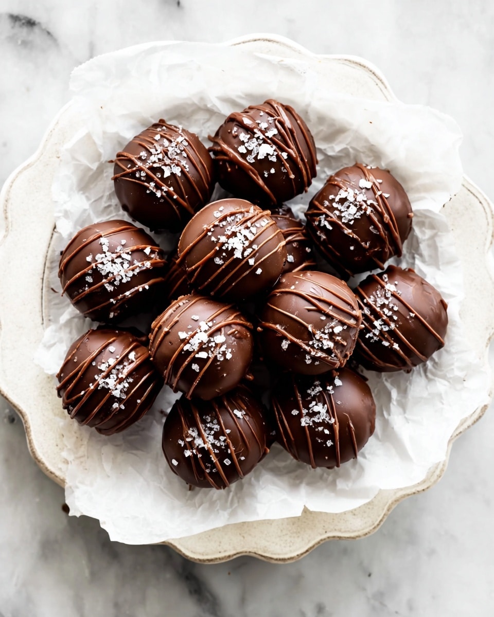 A white scalloped plate holds a layer of crumpled white parchment paper, on which sit about fifteen round, smooth chocolate truffles. Each truffle is covered in a glossy dark chocolate shell with thin, darker chocolate drizzle lines crossing the top, and sprinkled with coarse white sea salt flakes. The truffles are neatly clustered together, showcasing their shiny and textured surfaces on a white marbled background. photo taken with an iphone --ar 4:5 --v 7