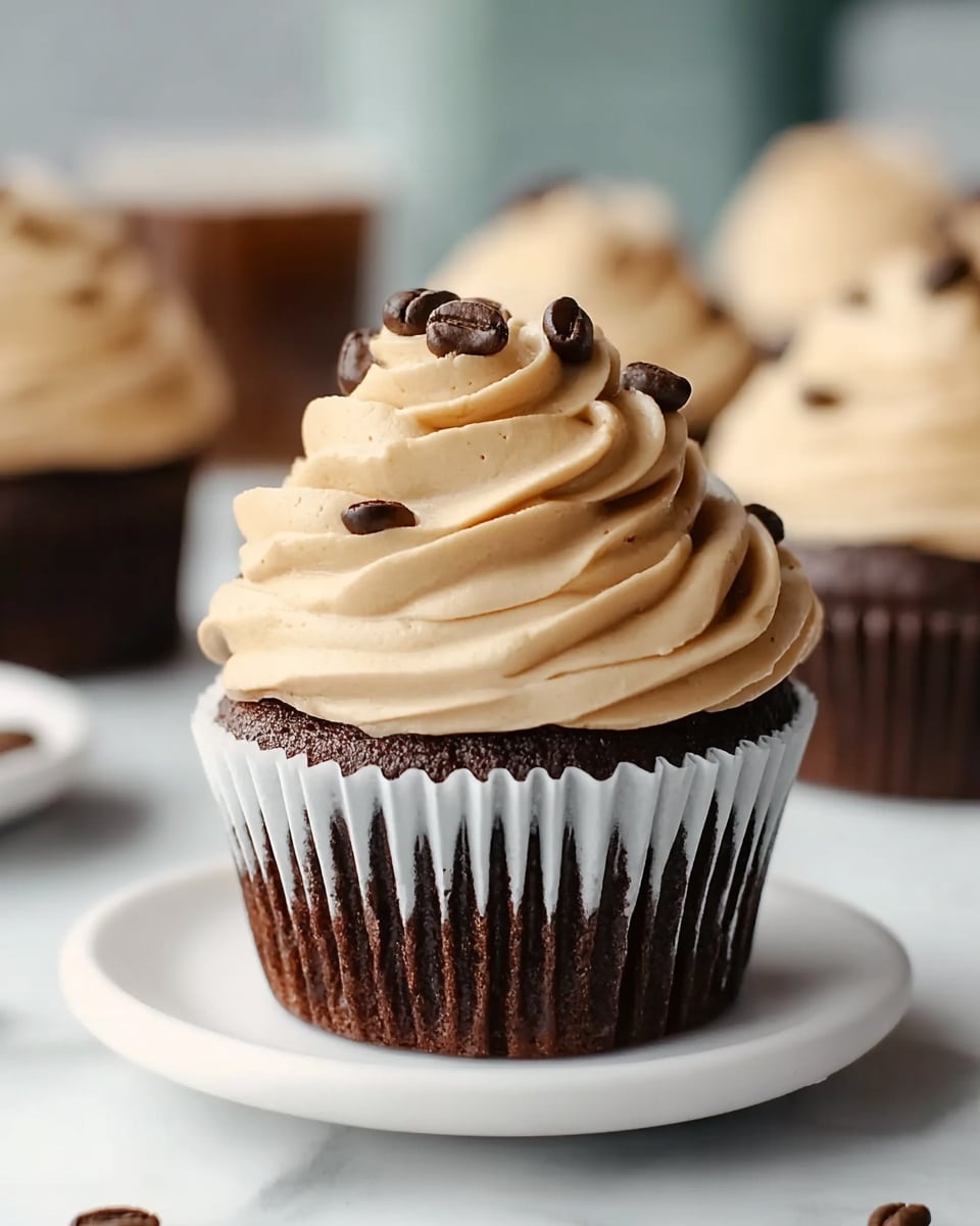 A close-up of a chocolate cupcake with a dark brown base, wrapped in a white paper liner. On top, there is a thick layer of light beige frosting swirled in three smooth, round layers with a soft, creamy texture. Small dark coffee beans are scattered on the frosting, adding contrast and detail. The cupcake sits on a clean white plate, set against a soft white marbled surface. In the blurred background, there are more cupcakes with the same frosting and coffee bean decoration. photo taken with an iphone --ar 4:5 --v 7
