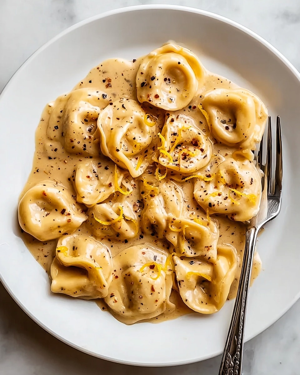 A white plate filled with a creamy sauce-covered tortellini pasta, showing about two layers of plump, ring-shaped tortellinis in a rich beige sauce. The sauce has a smooth texture with visible small specks of black pepper and some thin yellow strands mixed in. A shiny silver fork rests on the right edge of the plate. The plate is placed on a white marbled surface. photo taken with an iphone --ar 4:5 --v 7