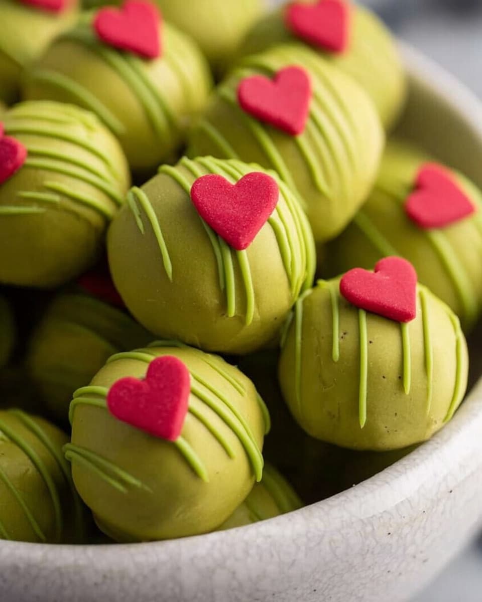 A close-up view of many round, green truffles arranged closely in a white bowl with a subtle texture. Each truffle is smooth and shiny with a bright olive green coating. Some truffles have thin green drizzle lines on their surface, while others are plain. On the top of every truffle, there is a small, flat, red heart-shaped decoration. The background is a soft, white marbled texture, making the green and red colors stand out clearly. Photo taken with an iphone --ar 4:5 --v 7