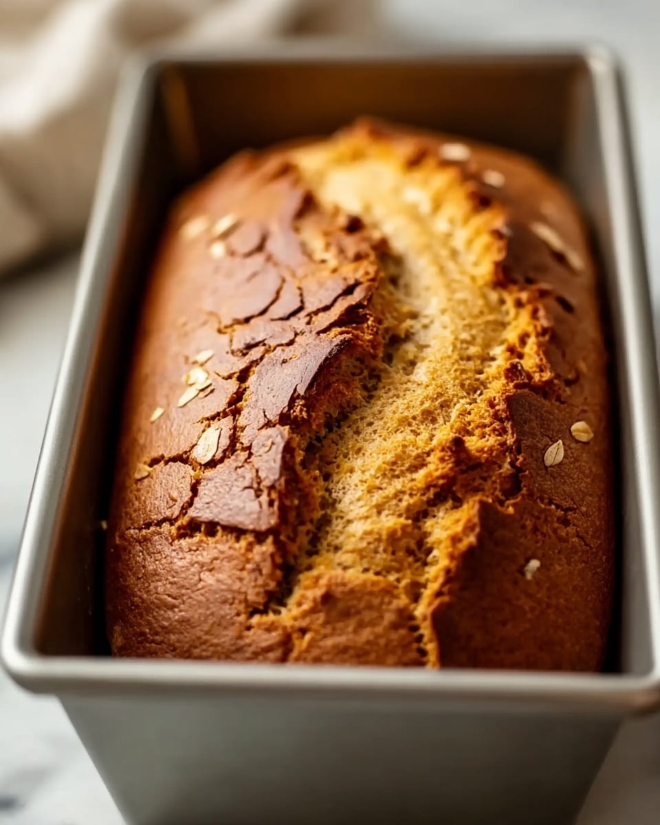 A close-up image of a freshly baked loaf of bread inside a silver metal baking pan. The bread has a cracked golden-brown crust on top with a deep central split revealing a lighter, softer inside texture. The edges of the loaf are slightly darker and the surface shows uneven cracks and rough texture. The pan rests on a white marbled surface with soft natural light coming from the left side, creating gentle shadows. photo taken with an iphone --ar 4:5 --v 7