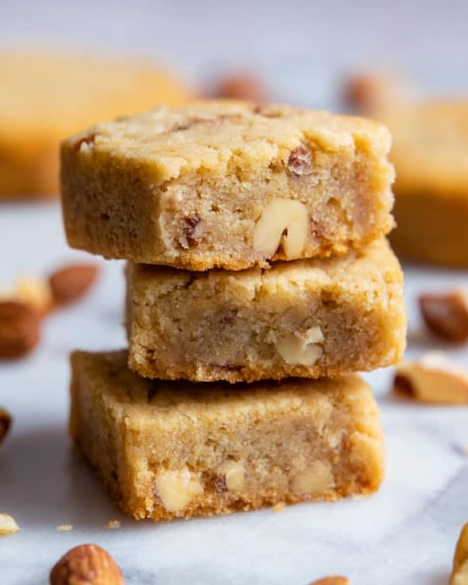 The image shows three square-shaped cookies stacked on top of each other on a white marbled surface. The cookies are light brown with a crumbly texture and small visible walnut pieces embedded throughout. Around the stack, some whole walnuts and blurred background elements are visible, adding a warm, cozy feeling to the scene. The lighting is soft and natural, highlighting the texture of the cookies. Photo taken with an iphone --ar 4:5 --v 7