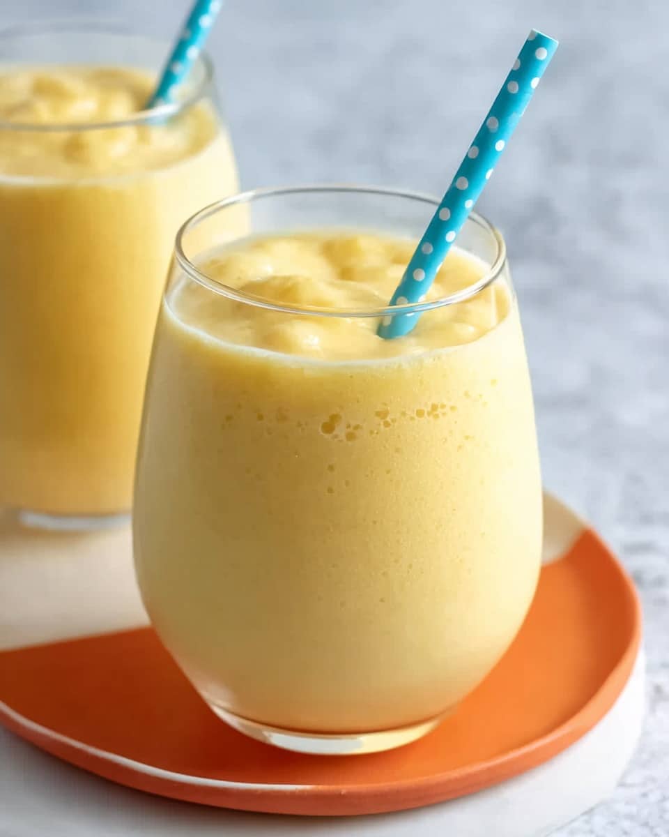 The image shows a small clear glass filled with a thick, pale yellow smoothie that has a smooth, creamy texture with slight bubbles on the surface. A light blue straw with white polka dots is dipped into the drink, angled slightly toward the viewer. The glass sits on a white plate with an orange smaller plate inside it, all placed on a white marbled surface. In the background, another glass of the same smoothie is partially visible, along with another straw of the same design lying flat. The overall look is fresh and inviting. photo taken with an iphone --ar 4:5 --v 7