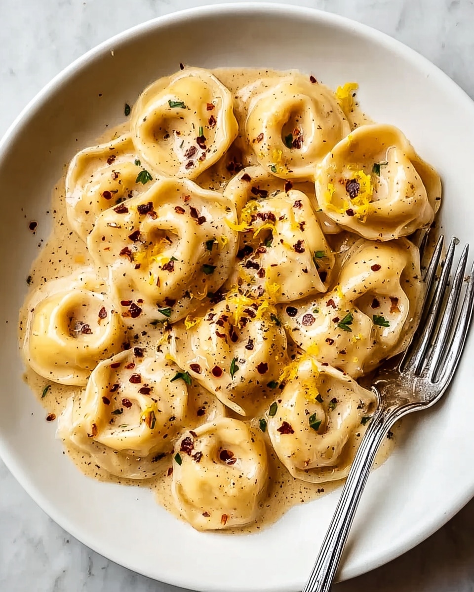 The image shows a white plate filled with about two layers of tortellini pasta coated in a creamy beige sauce. The sauce looks smooth and thick, covering the pasta well. There are small pieces of yellow zest mixed into the sauce, along with black pepper and red chili flakes sprinkled on top, adding specks of black and red. A shiny silver fork rests on the right side of the plate, partially touching the pasta. The plate is placed on a white marbled surface. photo taken with an iphone --ar 4:5 --v 7