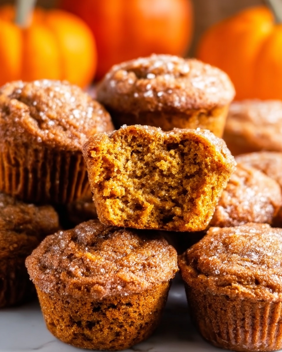 A close-up view of a group of golden brown muffins, with one muffin in the center placed on top of another, partly eaten to show its soft, moist inside texture with small air holes. The muffins have a slightly rough top sprinkled with sugar, giving a grainy look. They are lined in white paper cupcake liners, and a few small orange pumpkins are blurred in the white marbled background behind them. photo taken with an iphone --ar 4:5 --v 7