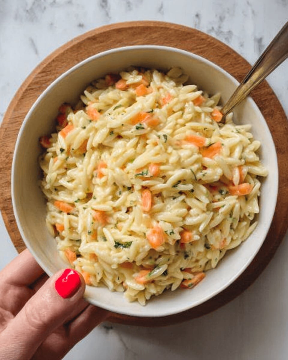 A close-up view of a white bowl filled with creamy orzo pasta mixed with small, finely chopped orange carrot pieces and tiny green herbs evenly spread throughout. The texture looks soft and slightly glossy, indicating a rich sauce coating the orzo. A woman's hand with red nail polish is holding the bowl from the side, and the background shows a circular wooden surface beneath the bowl and a white marbled texture around it. photo taken with an iphone --ar 4:5 --v 7