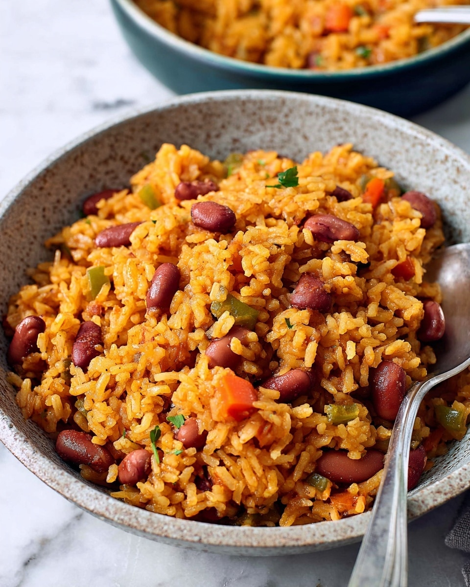 A close-up view of a large bowl filled with cooked rice mixed with red beans and diced tomatoes. The rice is yellow-orange and looks soft and fluffy, while the beans are whole and dark red. There are small pieces of green and red vegetables mixed in, adding contrast. The bowl itself is white with a speckled texture. A spoon rests inside the bowl, partially buried in the rice. All of this sits on a white marbled surface. Photo taken with an iphone --ar 4:5 --v 7