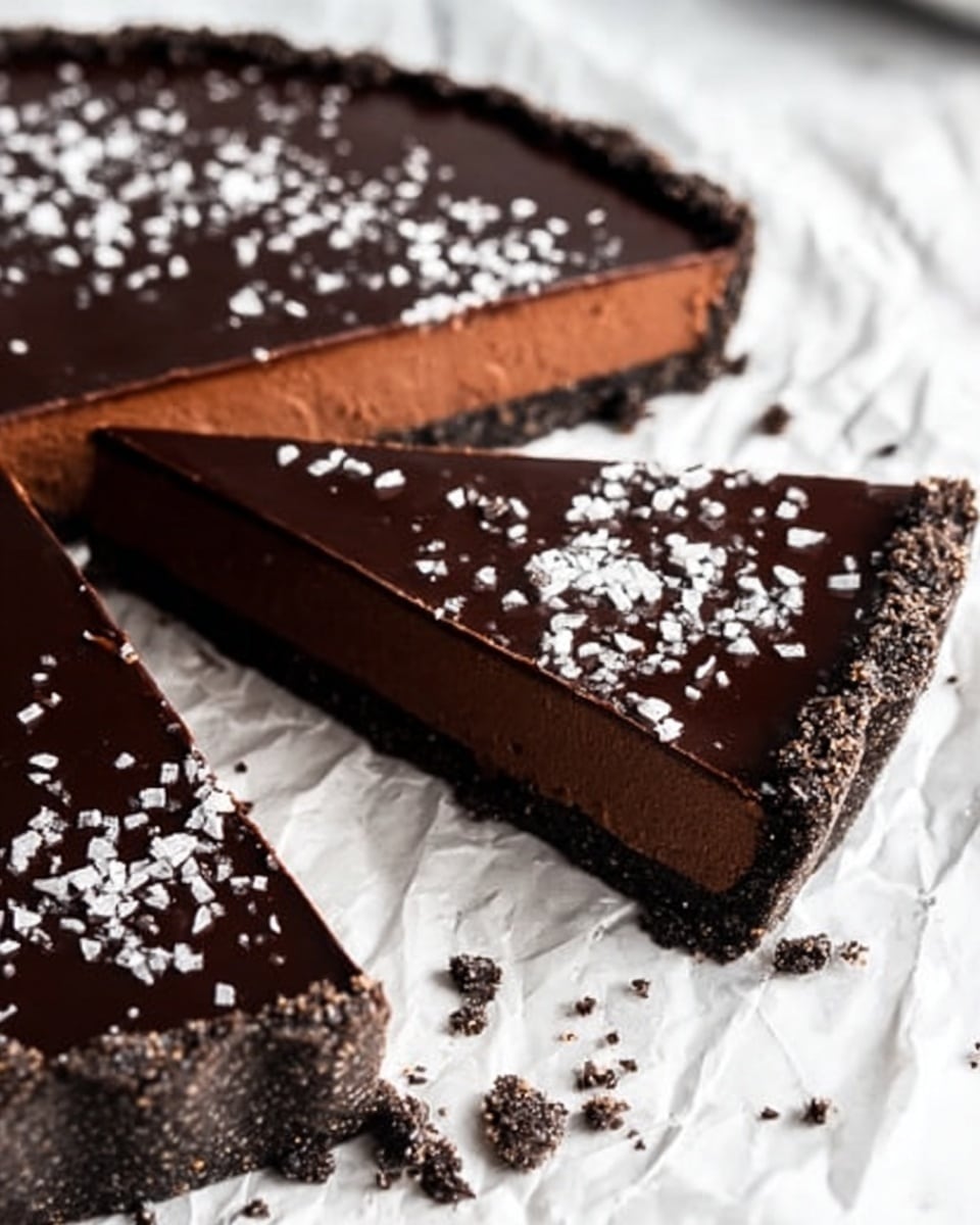 A close-up of a chocolate tart sliced into pieces, showing three distinct layers: a dark brown glossy chocolate ganache top, a middle layer of smooth milk chocolate mousse in a lighter brown shade, and a base of dark chocolate crumb crust that looks slightly rough and thick. The tart is on white marbled paper sprinkled with some dark chocolate crumbs and white flakes, creating a contrast with the smooth chocolate layers. The light shines softly on the ganache, highlighting its shine and the texture of the other layers. photo taken with an iphone --ar 4:5 --v 7