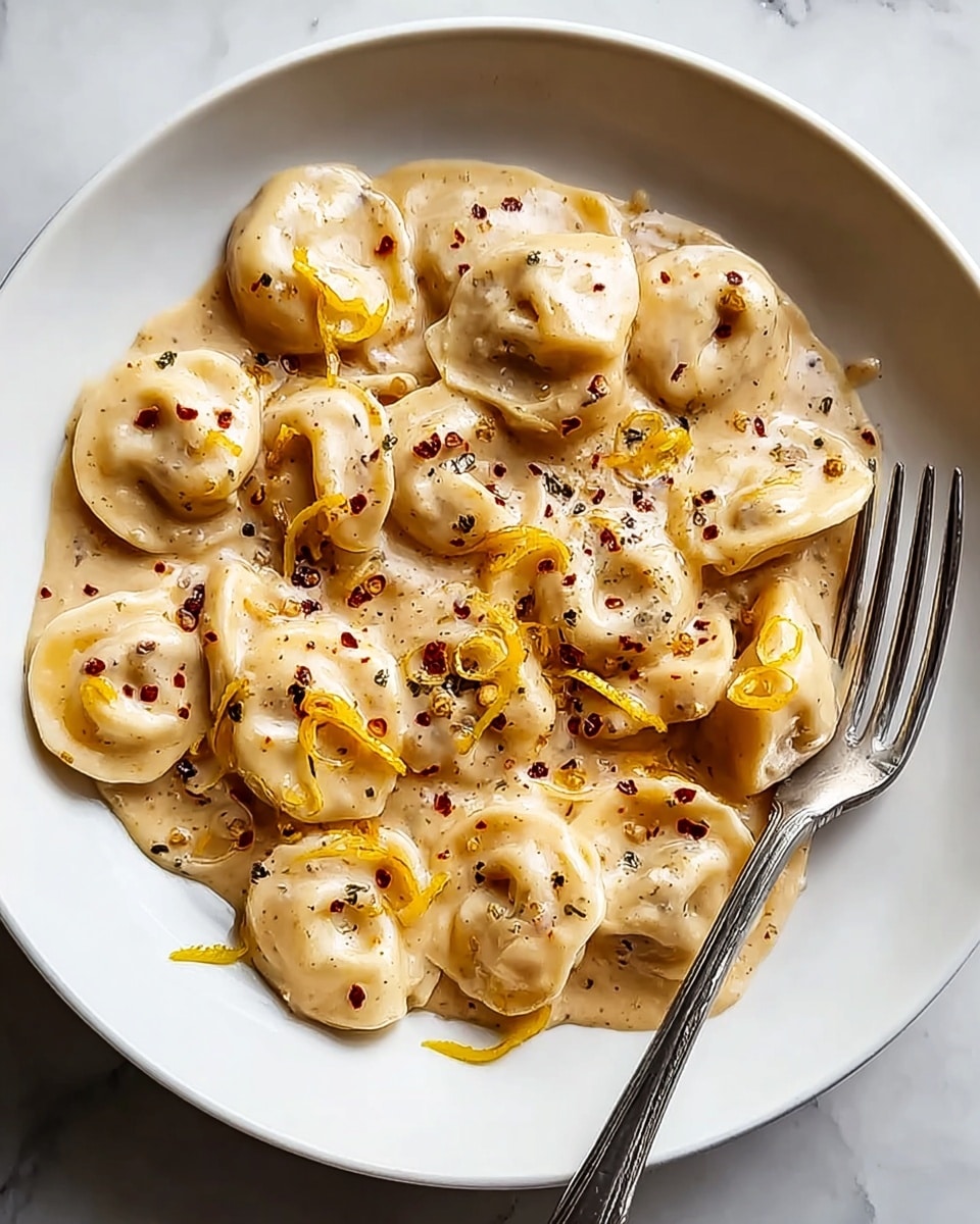 A white plate holds a creamy pasta dish with round, stuffed tortellini pieces covered in a thick, beige sauce. The sauce has a smooth texture with small black and red pepper flakes scattered on top. Thin strips of yellow zest or vegetable add color contrast throughout the dish. A silver fork rests on the right side of the plate. The meal sits on a white marbled surface. Photo taken with an iphone --ar 4:5 --v 7