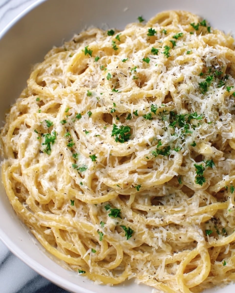 A close-up top view of a white plate filled with creamy spaghetti pasta, mixed evenly with a light beige sauce that has a smooth and rich texture. The pasta strands are twisted and layered throughout the plate, with small green herb bits sprinkled on top for a pop of color. Tiny shreds of pale grated cheese are scattered over the dish, adding texture and detail. The plate sits on a white marbled surface. photo taken with an iphone --ar 4:5 --v 7