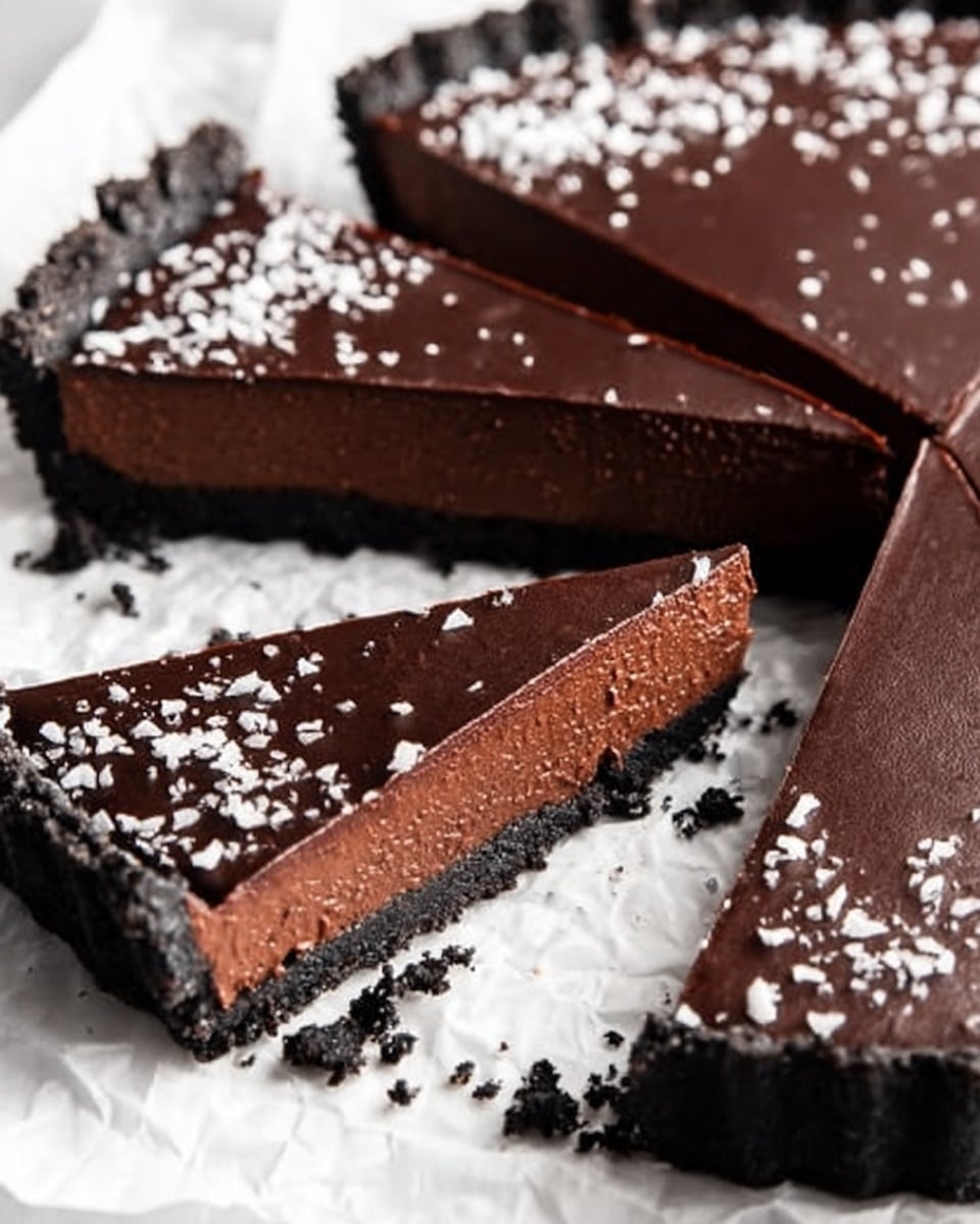 The image shows a close-up of a chocolate tart with four visible slices, placed on crumpled white paper on a white marbled surface. The tart has three layers: a thick, dark crumbly crust on the bottom, a dense and smooth medium brown chocolate filling in the middle, and a glossy, dark chocolate ganache layer on top sprinkled with white flakes. One slice is slightly pulled out, showing the clean edges and smooth texture of the layers. There is some crumbly crust scattered around the tart. photo taken with an iphone --ar 4:5 --v 7