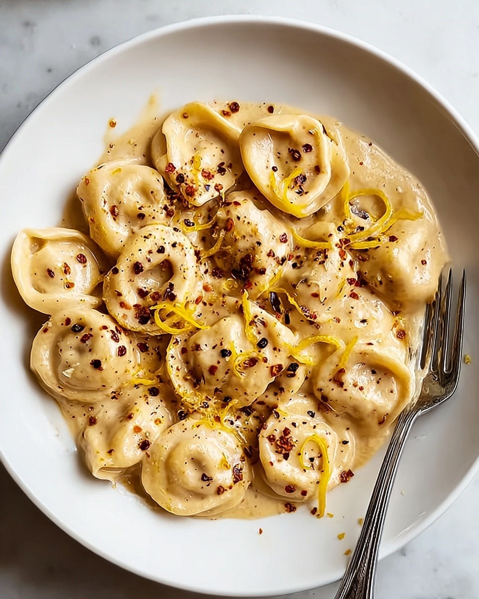 A close-up view of a white plate filled with about two layers of tortellini pasta coated in a creamy light beige sauce. The tortellini pieces are folded into ring shapes, visible in the top soft layer, with a smooth and slightly shiny texture. Thin strands of orange zest and small black specks of ground pepper are sprinkled evenly across the sauce. A silver fork rests on the right side of the plate, partially covered by the sauce, all placed on a white marbled surface. Photo taken with an iphone --ar 4:5 --v 7