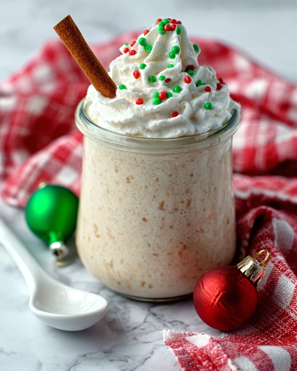The image shows a clear glass jar filled to the top with a thick, creamy beige drink that has a smooth texture and small visible bits inside. On top of the drink is a large swirl of white whipped cream decorated with small red and green round sprinkles. A cinnamon stick is placed standing upright in the whipped cream, slightly leaning to one side. The jar is on a white marbled surface with a red and white checkered cloth underneath. Nearby, a white ceramic spoon and two shiny Christmas ornaments, one green and one red, are placed. Photo taken with an iphone --ar 4:5 --v 7