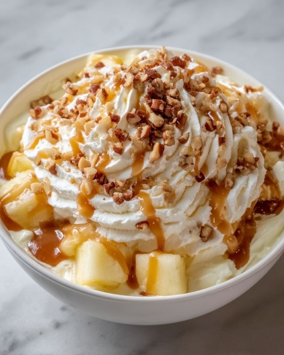 A white bowl filled with a creamy dessert showing three main layers: the bottom layer is small, soft, pale yellow apple pieces; the middle layer is smooth white cream covering the apples; the top layer is golden caramel sauce drizzled over the cream, topped with a pile of crunchy, light brown granola pieces. The bowl sits on a white marbled surface. Photo taken with an iphone --ar 4:5 --v 7