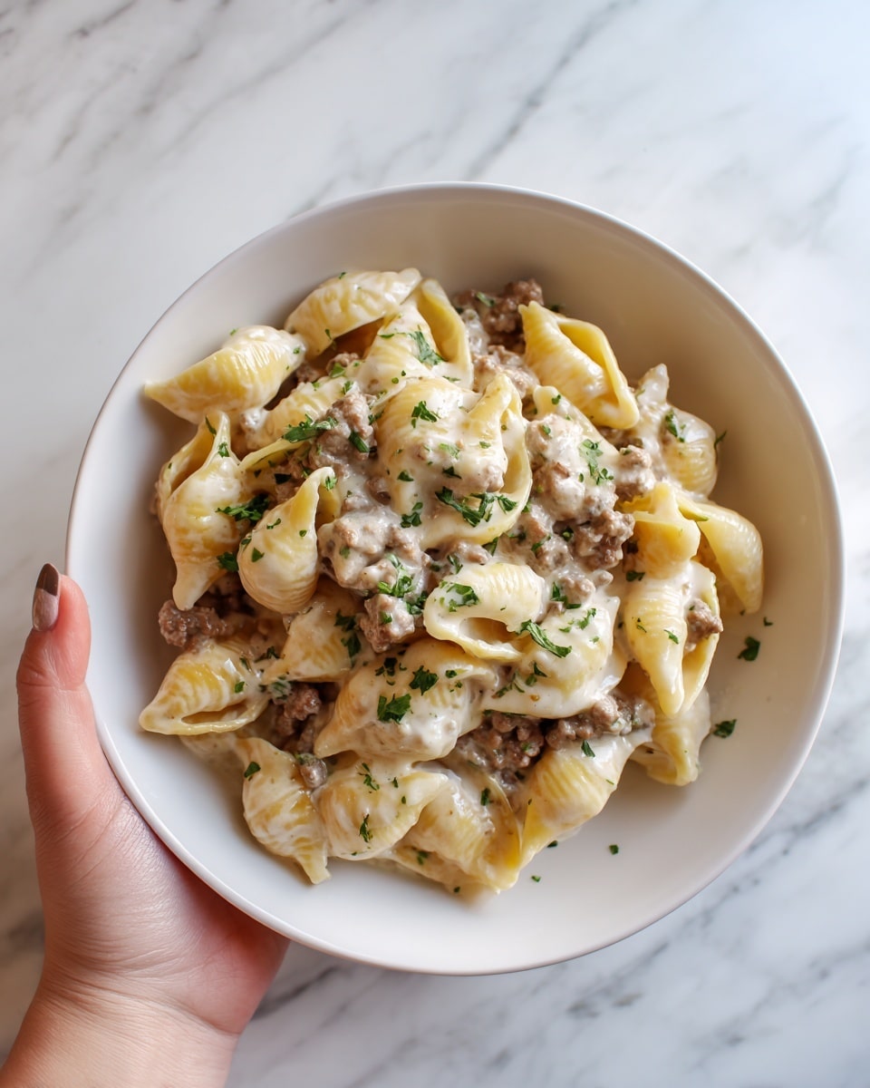 The image shows a bowl of creamy pasta with three visible layers: the bottom layer is white pasta shells with a smooth texture, the middle layer is a creamy white sauce that coats the pasta evenly, and the top layer is browned ground beef mixed with small green herb pieces sprinkled throughout. The bowl is white and sits on a white marbled surface, with a slightly blurred green garnish visible in the background. The dish looks rich and well blended, with a soft and hearty feel. Photo taken with an iphone --ar 4:5 --v 7
