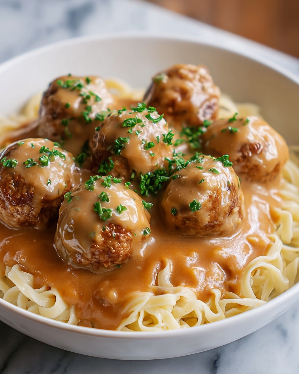 A white bowl filled with a base layer of light beige cooked noodles, soft in texture and slightly curled. On top, there are six brown meatballs that are moist and coated with a light brown, creamy gravy sauce. Small green chopped herbs are sprinkled over the meatballs and sauce, adding a fresh touch. The bowl sits on a white marbled surface, and the photo is taken close-up to show the textures and colors clearly. photo taken with an iphone --ar 4:5 --v 7