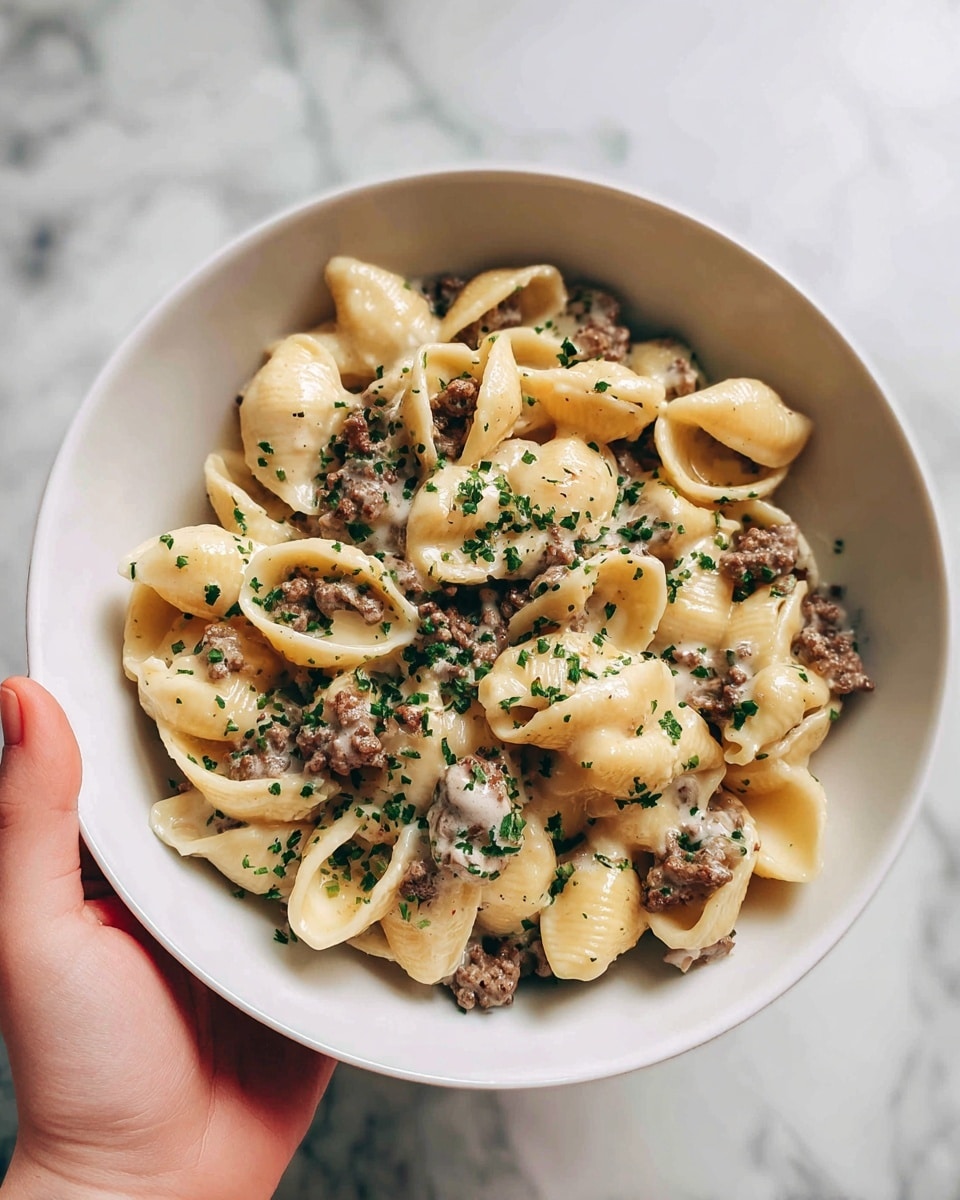 A white bowl filled with creamy pasta shell shapes mixed with ground beef pieces, small green chopped herbs sprinkled on top. The sauce looks smooth and white with a slightly glossy texture, coating the pasta evenly. There is a woman's hand holding the bowl on the side, with a white marbled surface in the background. Photo taken with an iphone --ar 4:5 --v 7