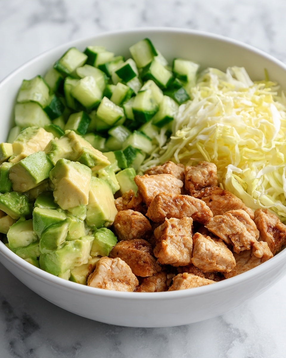 A white bowl with four distinct sections filled with food on a white marbled surface. One section shows green diced avocado pieces with a soft texture. Next to it, there is a pile of bright green chopped cucumbers with a fresh look. Another part contains cooked chicken pieces with a slightly crispy brown surface. The last section is filled with a layer of shredded light yellow cabbage or lettuce. photo taken with an iphone --ar 4:5 --v 7