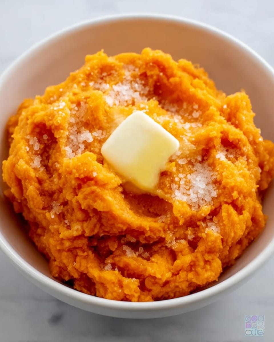 In the image, there is a white bowl filled with bright orange mashed sweet potatoes that have a rough, chunky texture. On top of the sweet potatoes is a melting pat of light yellow butter creating a slight glossy shine, and around it, small sprinkles of white granulated salt or sugar can be seen. The bowl sits on a white marbled surface, giving a clean and fresh look. Photo taken with an iphone --ar 4:5 --v 7
