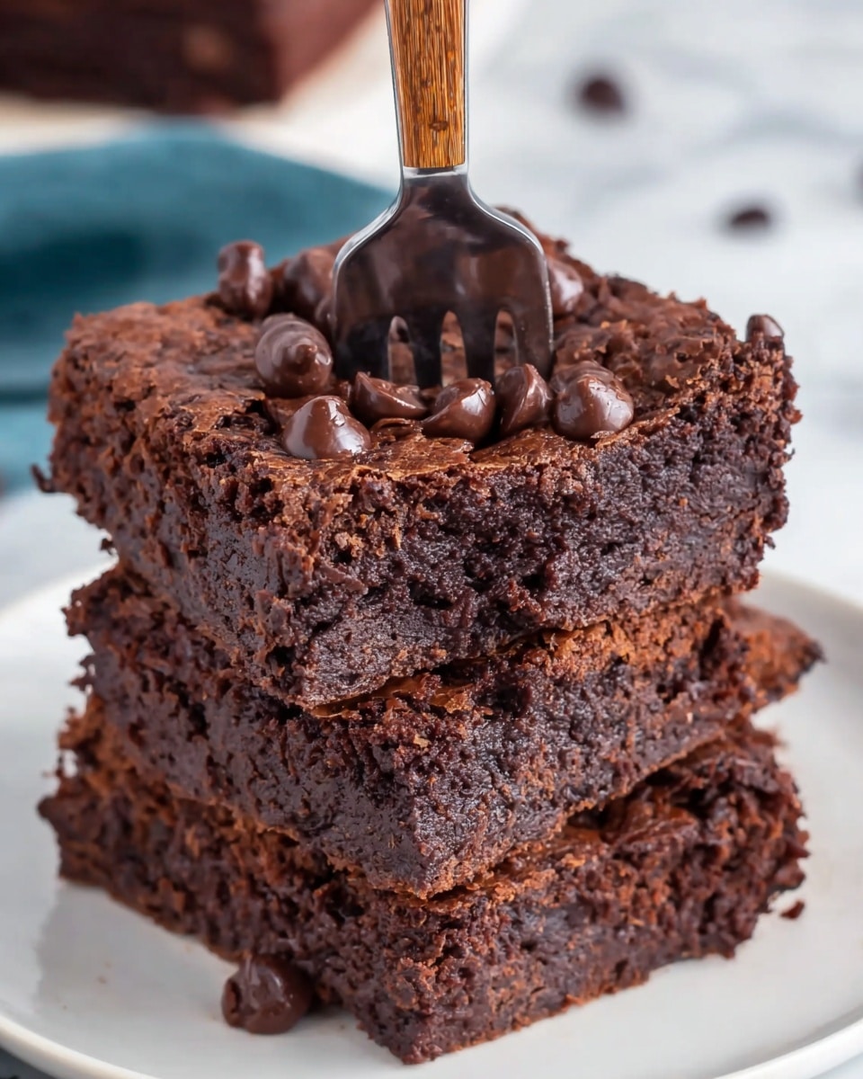 A close-up view of a stack of three thick, rich chocolate brownies on a white plate, each brownie showing a moist and dense texture with a slightly crumbly top layer sprinkled generously with chocolate chips and small chocolate chunks. The fork is pressed into the top brownie, revealing its fudgy inside that looks soft and gooey. The top layer is darker and shiny with melted chocolate bits, while the middle and bottom layers are deep brown with a dense, cake-like texture. The background is a white marbled texture, softly out of focus, emphasizing the chocolatey treats. Photo taken with an iphone --ar 4:5 --v 7