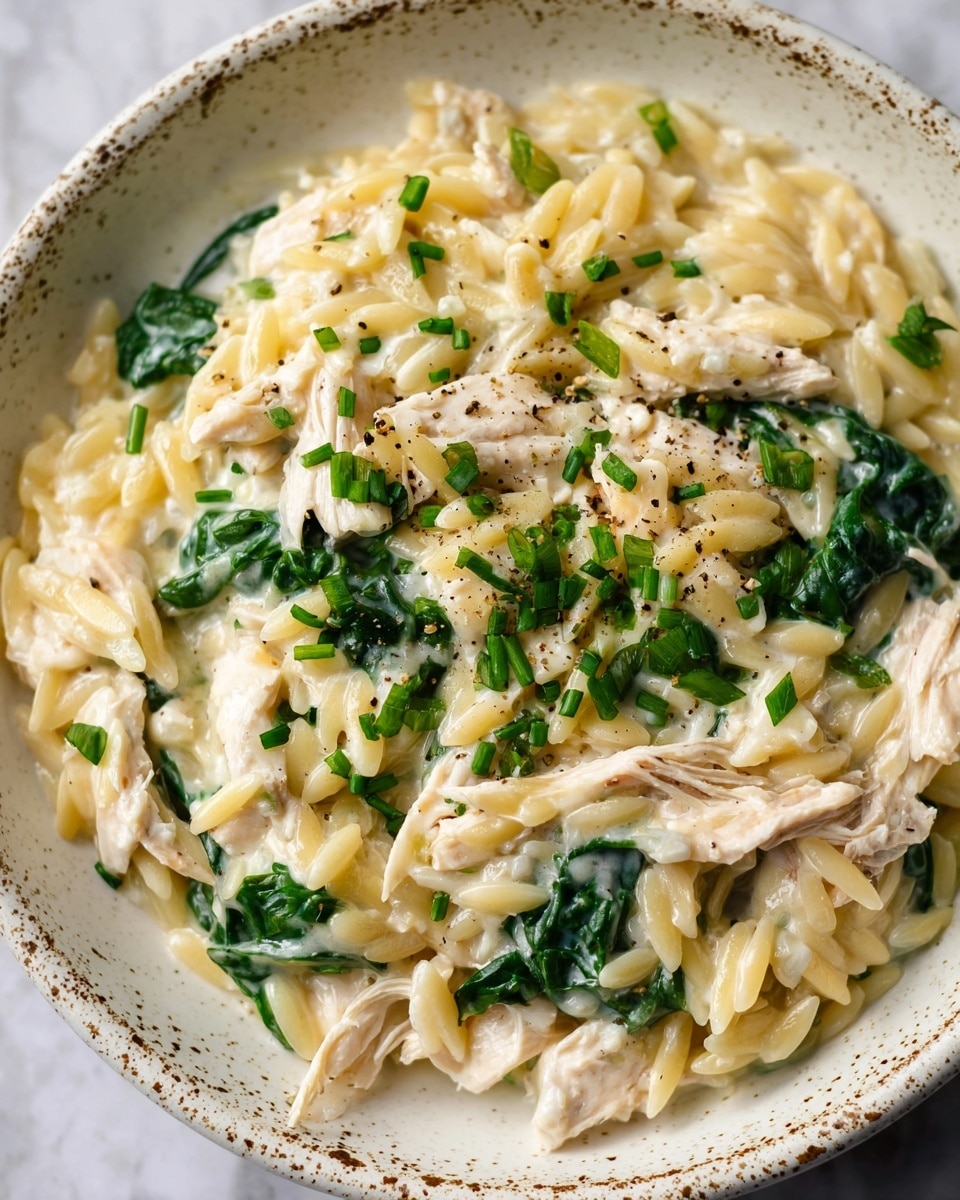 The image shows a close-up of a creamy orzo pasta dish served in a white bowl with a speckled pattern. The dish has multiple layers: small orzo pasta with a pale yellow color, tender white shredded chicken pieces mixed throughout, and vibrant dark green spinach leaves nestled in the creamy sauce. The creamy sauce is light and smooth, coating all the ingredients. Finely chopped fresh green herbs are sprinkled evenly over the top, adding a fresh contrast. There is a slight shine on the surface from the sauce, and hints of black pepper are visible for seasoning. The bowl rests on a white marbled surface. photo taken with an iphone --ar 4:5 --v 7