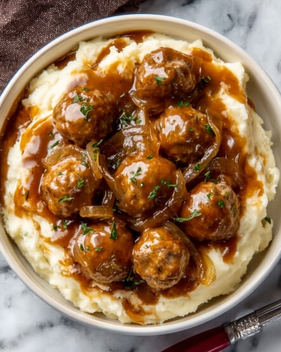 A round white bowl filled with a creamy, smooth base layer of mashed potatoes that is off-white in color. On top, there is a thick layer of brown meatballs covered in a glossy brown onion gravy with visible soft, cooked onion slices mixed in. Small green bits, likely herbs, are scattered lightly on the meatball and gravy layer for garnish. The bowl is set on a white marbled surface with a silver fork beside it. Photo taken with an iphone --ar 4:5 --v 7