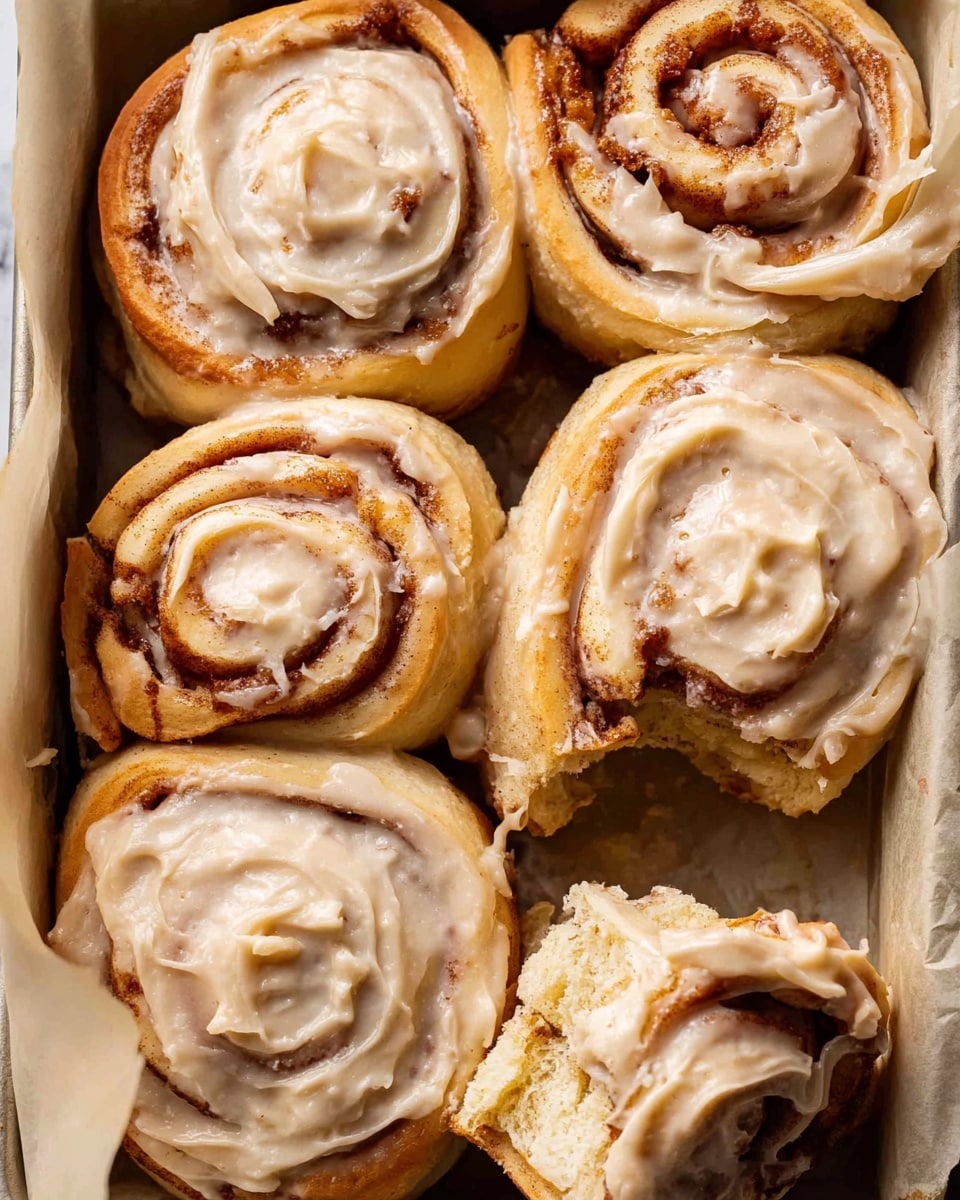 A close-up view of six cinnamon rolls in a baking pan lined with parchment paper, five of the rolls are whole and one is partially torn open showing its soft, layered dough with cinnamon swirls inside. The cinnamon rolls are topped with a creamy, light beige frosting spread thickly over the spiraled tops, allowing the cinnamon layers to peek through. The dough’s outside is a light golden-brown, and the rolls are tightly arranged, emphasizing their soft, fluffy texture against the lined pan. The background is a white marbled texture. photo taken with an iphone --ar 4:5 --v 7