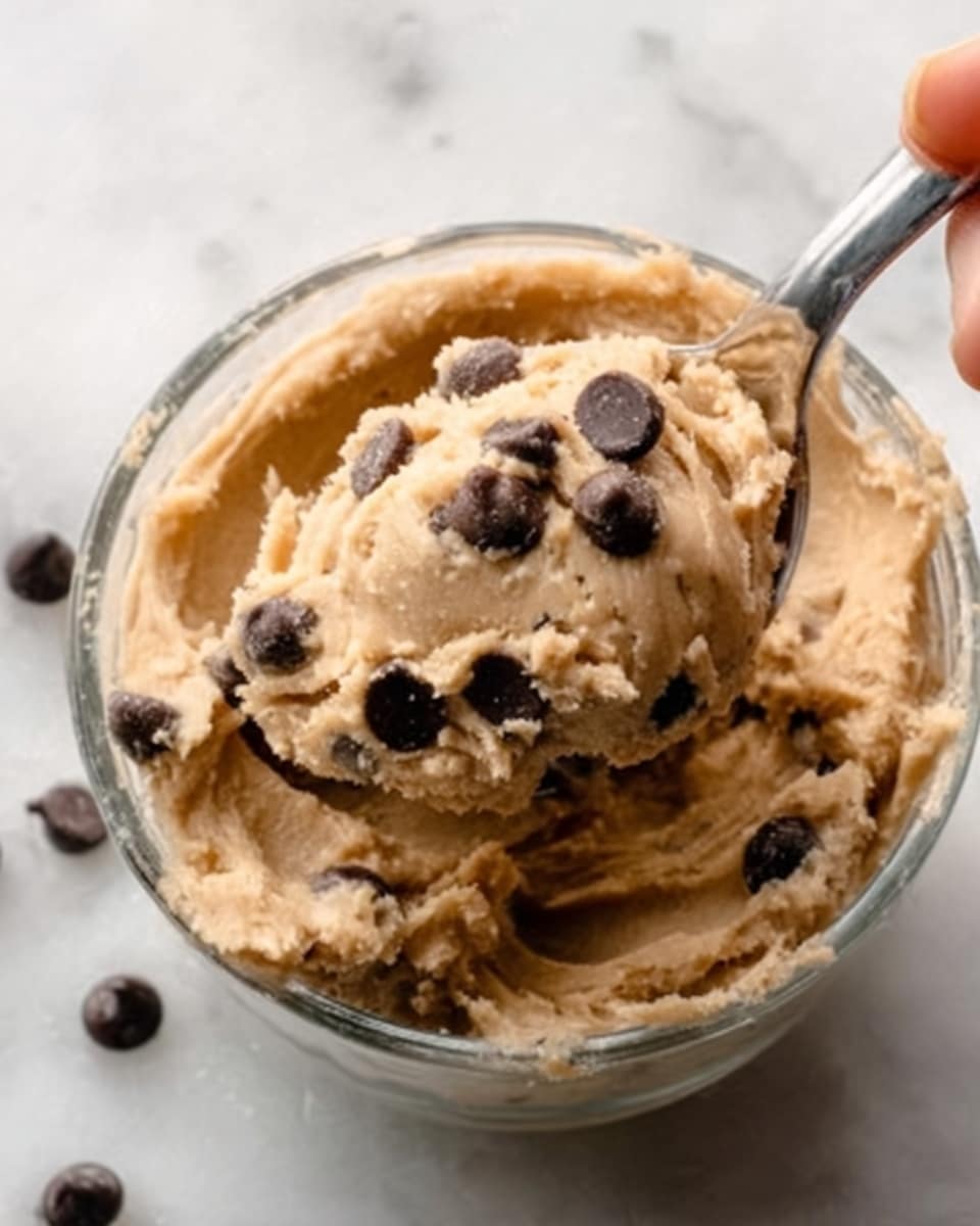 A close-up image of a small glass bowl filled with creamy, light brown cookie dough mixed with several glossy dark brown chocolate chips. The cookie dough has a smooth texture with small bumps where the chocolate chips are embedded. One scoop of the dough is slightly raised and rounded at the center, showing the thick consistency. A woman's hand is using a silver spoon to lift some of the dough from the bowl. The bowl sits on a white marbled surface. Photo taken with an iphone --ar 4:5 --v 7
