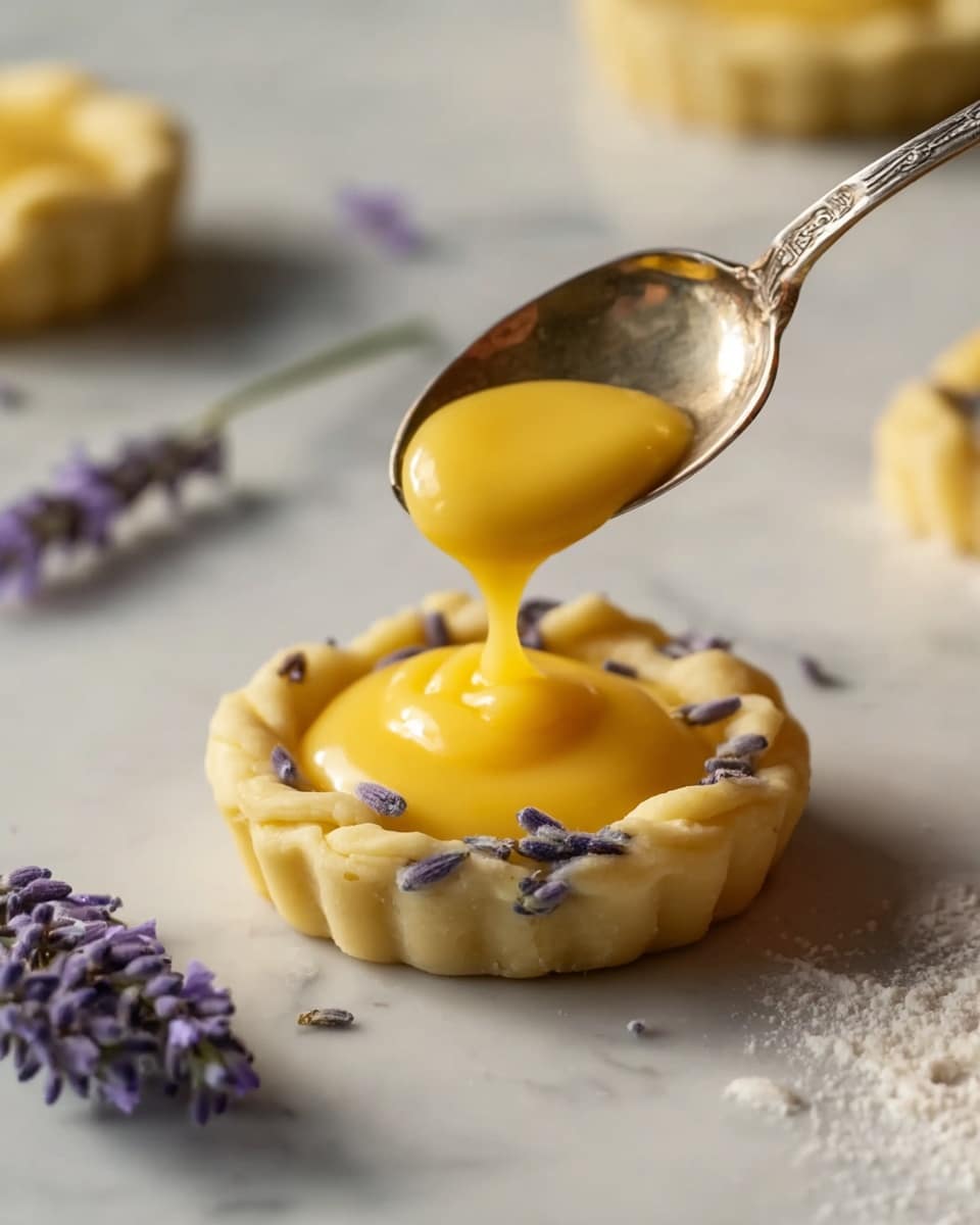 The image shows a close-up of a small tart with two layers. The bottom layer is a crinkled, pale yellow dough embedded with small purple lavender buds, shaped into a round base with slightly uneven edges. The top layer is a smooth, glossy, bright yellow custard filling sitting neatly inside the dough shell. A silver spoon, held above the tart, is adding more of the same glossy custard filling onto the top, creating a small mound. The tart rests on a white marbled surface scattered with some flour and a few sprigs of lavender nearby, adding a delicate touch to the scene. Photo taken with an iphone --ar 4:5 --v 7