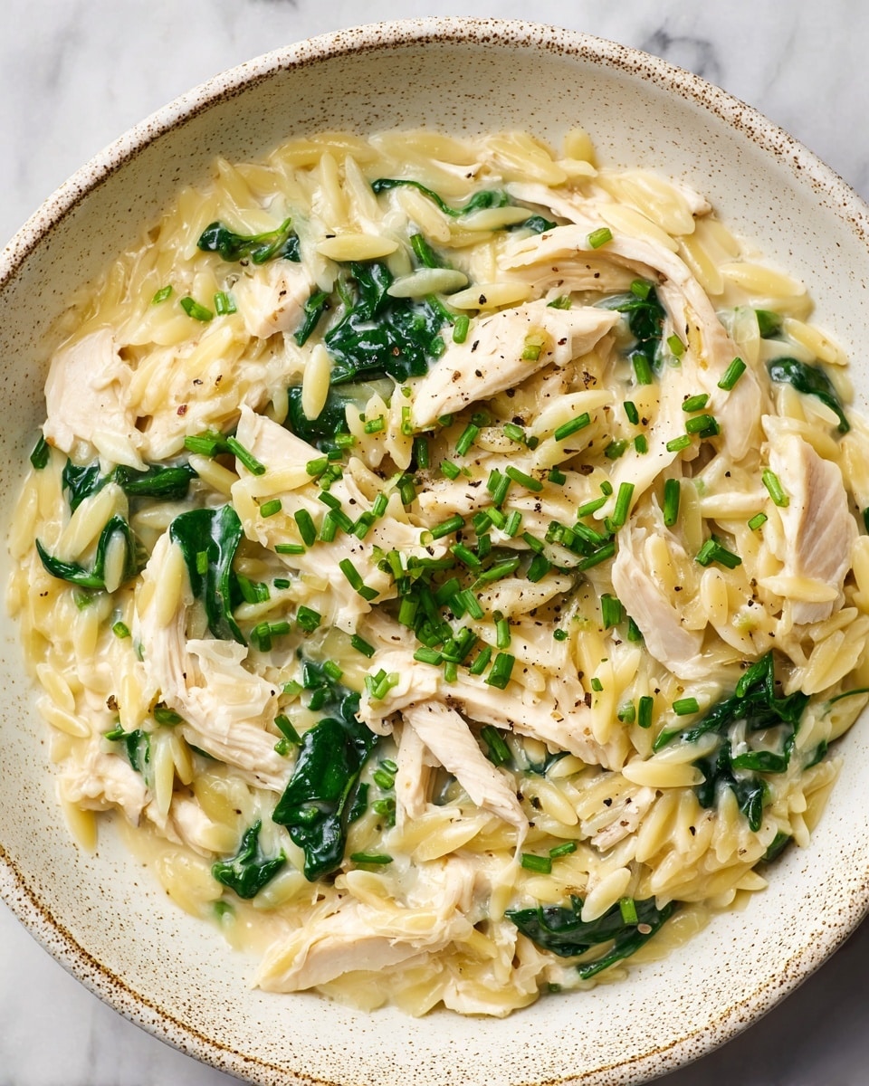 A close-up of a creamy orzo pasta dish served in a white bowl with subtle brown speckles. The dish has several layers starting with the orzo pasta, which is pale yellow and coated in a white creamy sauce. Mixed throughout are tender pieces of shredded white chicken and vibrant dark green spinach leaves, adding color contrast and texture. Small green chopped herbs, likely chives, are sprinkled on top, along with a light dash of ground black pepper. The dish sits on a white marbled textured surface. Photo taken with an iphone --ar 4:5 --v 7