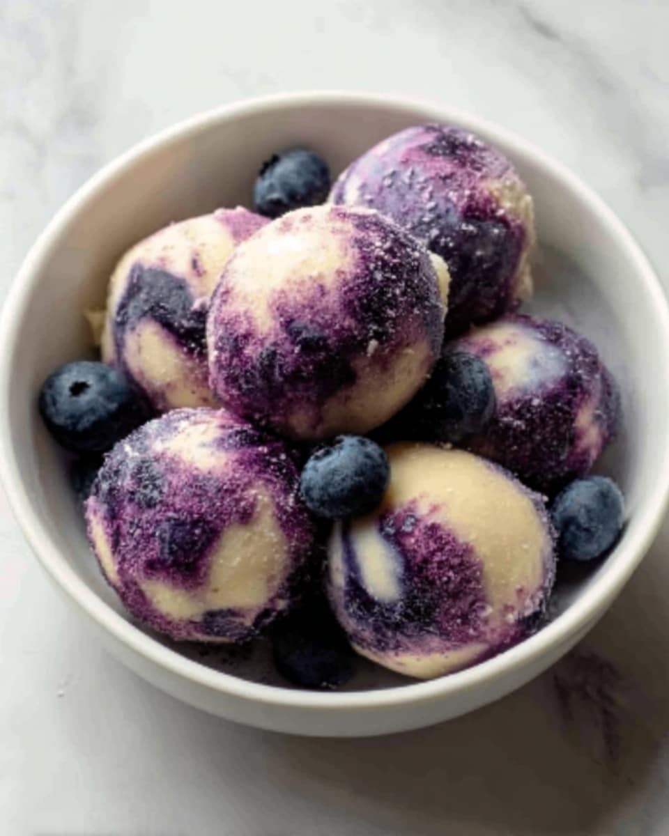 A white bowl filled with five round balls of dessert that are mainly light cream colored with swirls and patches of deep purple, giving a marbled look. The balls have a smooth but slightly bumpy texture, and some purple powder is lightly sprinkled on top of them. There are a few fresh blueberries arranged around the balls inside the bowl. The bowl is set on a surface with a white marbled texture. Photo taken with an iphone --ar 4:5 --v 7