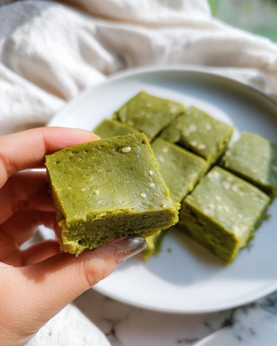 A close-up of a green dessert cut into nine square pieces on a white plate, placed on a white marbled surface. The dessert has a smooth and slightly cracked top layer showing its dense and moist texture. One square piece is held by a woman's hand above the plate, revealing the dessert's thick, soft, and crumbly middle layer with a bright green color. The background includes a soft, light-colored cloth. photo taken with an iphone --ar 4:5 --v 7