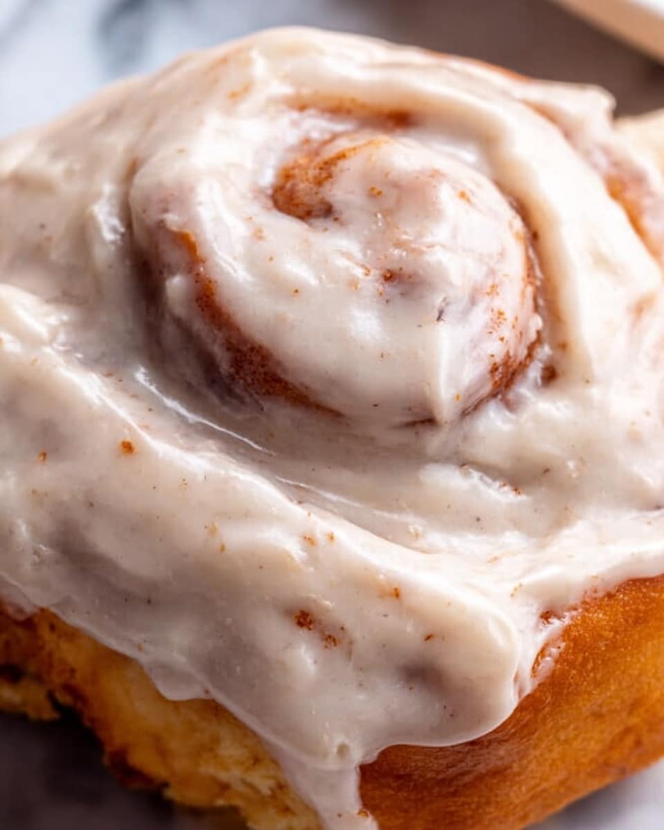 A close-up view of a cinnamon roll covered in thick, creamy white icing with a slightly speckled texture from cinnamon or spices. The roll shows a visible swirl of golden-brown dough peeking through the smooth, glossy icing, which covers the whole top evenly and spills down the sides. The background surface is a white marbled texture, enhancing the warm and soft look of the cinnamon roll. photo taken with an iphone --ar 4:5 --v 7